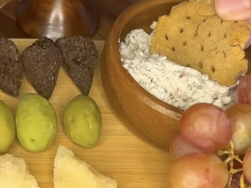 Wooden board with olives, cheese spread on perforated cracker, onions, and dried figs